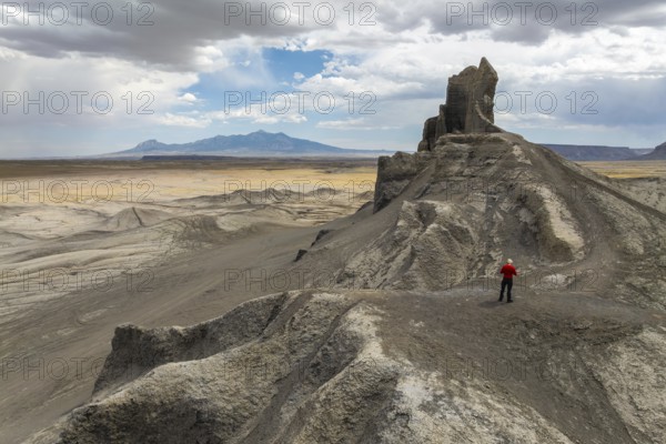 A person in a red jacket stands atop a rugged, rocky outcrop in a vast desert landscape in Hanskville, Utah. The dramatic geological formation contrasts with the expansive, barren surroundings