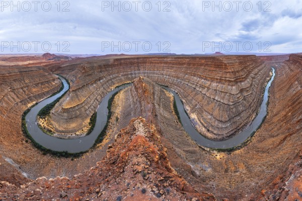 A breathtaking view of a winding river carving through a deep, red rock San Juan canyon, showcasing stunning geological formations beneath a vast, cloudy sky in a remote desert in Utah, USA