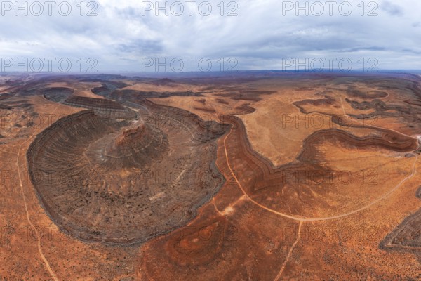 Aerial view of a vast desert landscape showcasing unique geological formations and winding paths in San Juan canyon. The red and brown hues contrast with the cloudy sky, highlighting natural beauty