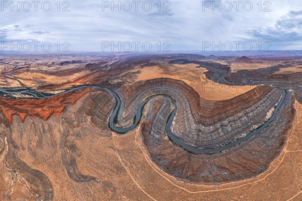 A stunning aerial view captures a meandering river cutting through the rugged San Juan canyon. Layers of red and brown rock form the dramatic landscape under a cloudy sky in Utah, USA