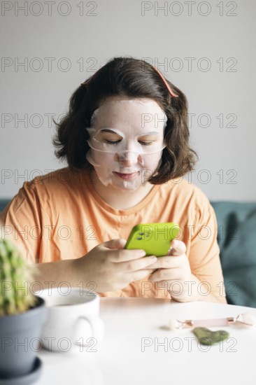 Woman at home, wearing facial beauty mask while browsing on her smartphone. A peaceful self-care routine captured indoors