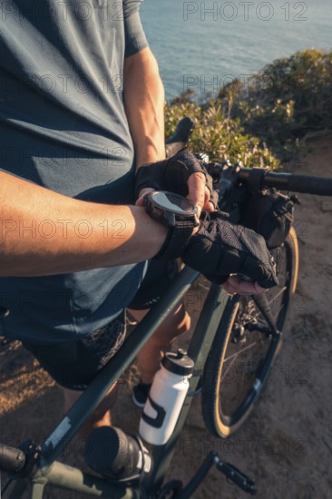 Cropped unrecognizable male cyclist leaning on his bicycle, looking at his watch, with a scenic ocean backdrop at Zahara de los Atunes, Cadiz