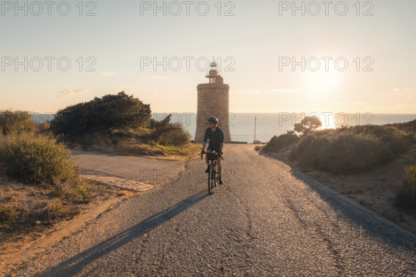 A man biking down a gravel road towards the historic Faro Camarinal lighthouse during sunset, with the sea in the background, located in Zahara de los Atunes, Cadiz