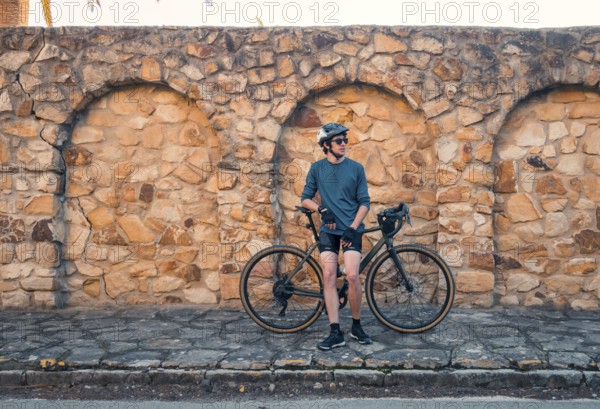 A young male cyclist stands next to his road bike, leaning against an aged stone wall in Zahara de los Atunes, Cadiz. The man is wearing sunglasses, a helmet, biking gear, and poses casually during a sunny day