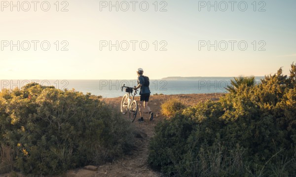 Back view of unrecognizable male cyclist standing with his bike, overlooking the ocean along a coastal trail near Faro Camarinal, Zahara de los Atunes, Cadiz during sunset
