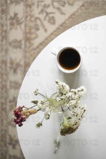 Top view of a tranquil setting featuring a bird's-eye view of a white cup filled with coffee beside a vase with dried flowers arranged loosely, set on a white circular table with a patterned background