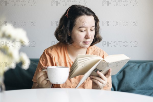 Young woman reads intently while holding a cup of coffee, comfortably seated on a sofa at home, embodying a peaceful domestic moment