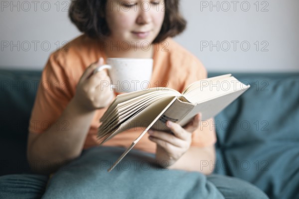 Cropped view of a woman sipping tea and reading a book, comfortably seated on a sofa