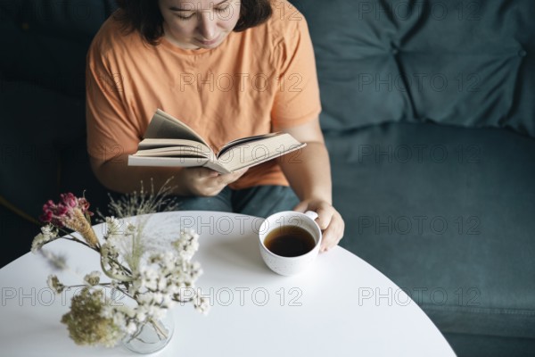 Focused woman reading a book and sipping coffee at a round white table, with a relaxed atmosphere at home, enhanced by soft, natural lighting and simple decor