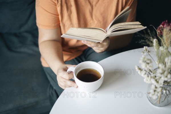 Cropped unrecognizable woman reading a book while holding a cup of coffee, seated comfortably at home