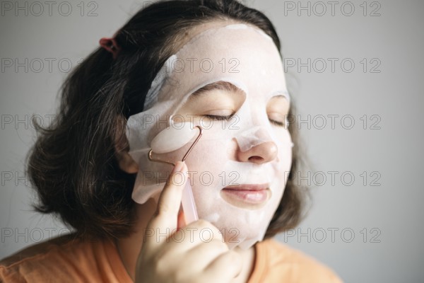 Image of a woman at home, using a facial pink quartz roller while wearing a sheet mask to enhance her skincare routine. Her eyes are closed, showing a moment of relaxation
