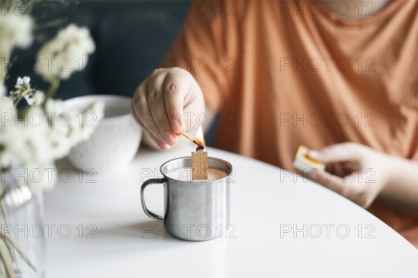 Cropped view of unrecognizable woman in a tangerine top lighting a candle inside a metal mug, fostering a serene home setting. A blurry vase with white flowers adds to the tranquil scene