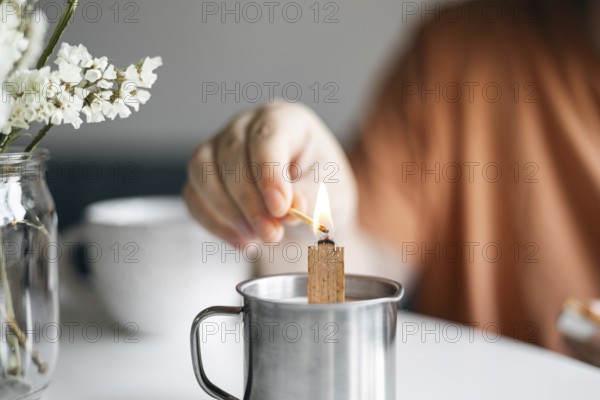 Cropped unrecognizable woman lights a candle placed in a metal mug, creating a cozy atmosphere at home. A vase with white flowers adds to the tranquil setting