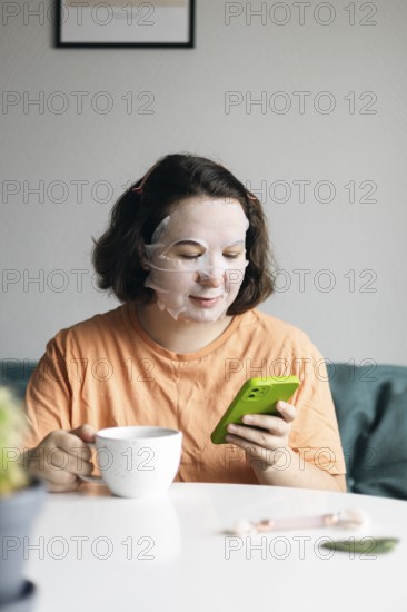 Woman with a facial mask resting at home while holding a cup of coffee and browsing on her smartphone, showcasing a moment of self-care and digital connectivity