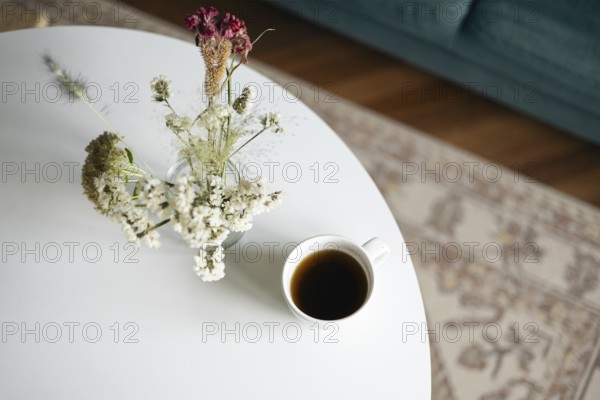 Top view of a serene home scene featuring a cup of black coffee and a vase of delicate dried flowers on a sleek white table, evoking a peaceful, restful atmosphere