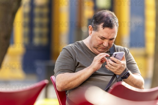 A blind man sitting on a red chair outdoors, using his smartphone. The background features muted urban tones and sunlight, creating a relaxed and focused atmosphere