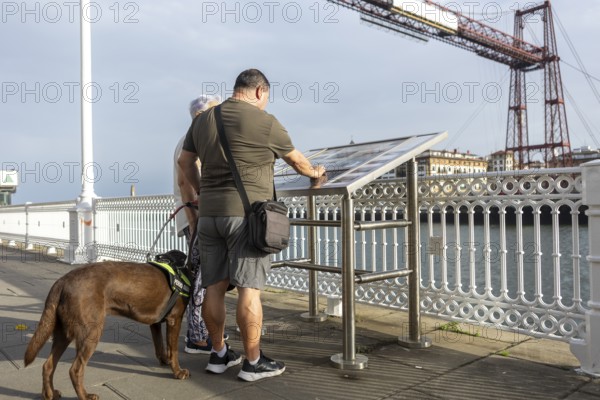A blind man with a guide dog read an information board on a bridge overlooking a scenic river and distant buildings. The bridge industrial design adds to the charm