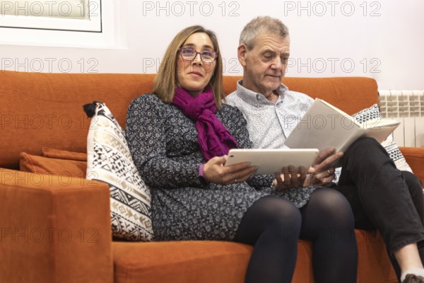 A couple with intellectual disabilities enjoys a evening indoors of a supervised home. The woman uses a tablet while the man reads a book. They sit on an orange sofa, creating a cozy atmosphere