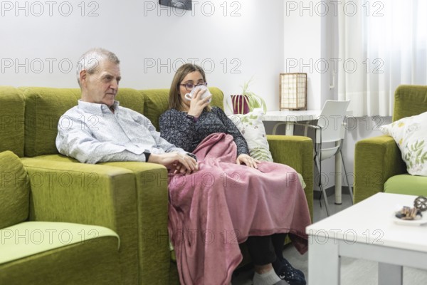 A couple with intellectual disabilities sit comfortably on a green sofa wrapped in a pink blanket. The woman enjoys a warm drink, creating a cozy and relaxing atmosphere at supervised home