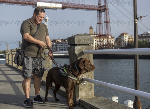 A blind man walks with his guide dog on a riverside path, showcasing companionship and accessibility. Urban landscape and architectural background add depth
