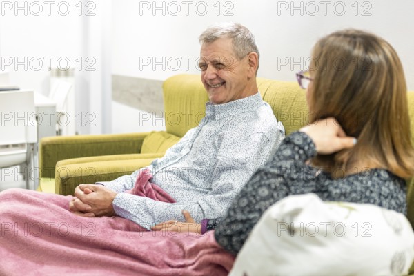 A couple with intellectual disabilities share a warm moment on a comfortable sofa, wrapped in a pink blanket. The bright setting and casual attire create a relaxed atmosphere at supervised home