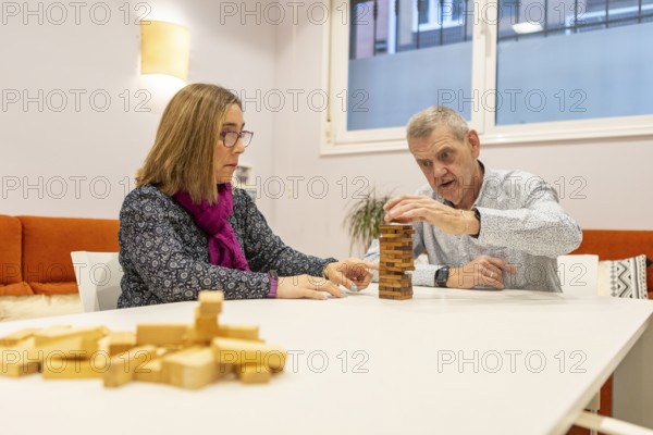 A couple with intellectual disabilities engaged in a game with wooden blocks on a table of a supervised home. A woman observes while a man carefully places a block. Brain exercises and patience