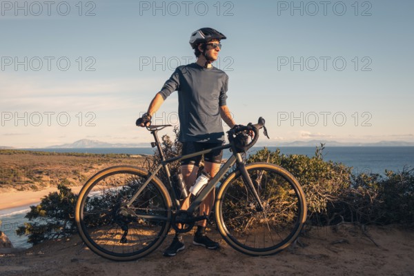 A male cyclist stands with his bike, overlooking the scenic Zahara de los Atunes, Cadiz during a sunset. The clear sky and coastal landscape provide a stunning backdrop
