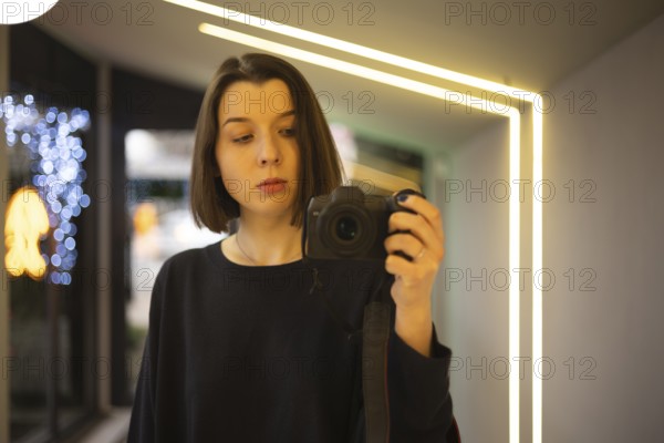 A woman capturing a self-portrait using a DSLR camera in a well-lit interior with neon accents and blurred festive lights. A blend of minimalism and modern style
