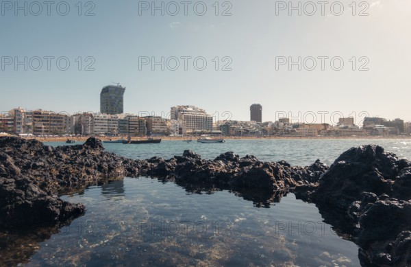 Tranquil cityscape view from volcanic rocks on a sunny day, showing modern buildings and calm waters in Las Palmas, Gran Canaria
