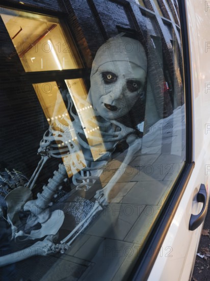 A haunting skeleton figure peers through a city window, its eerie reflection captured on the glass. The scene blends urban architecture with Halloween-style themes