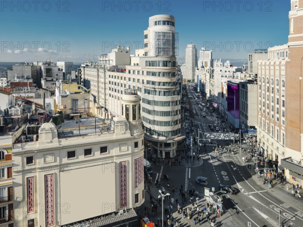 A bustling urban Madrid scene captures the vibrant life of Gran Via street flanked by historic buildings. Pedestrians and vehicles fill the road under a clear blue sky, showcasing city liveliness