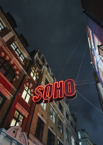 A vibrant neon Soho sign illuminates the street below, contrasting against a dark, moody London night sky. The iconic scene captures the lively spirit of the area