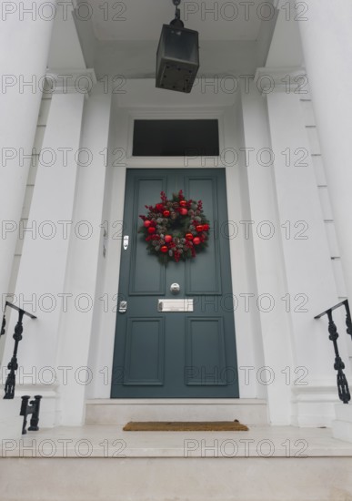 A stylish London doorway features a festive wreath with red ornaments, pinecones, and greenery against a teal door. White columns and iron railings add elegance