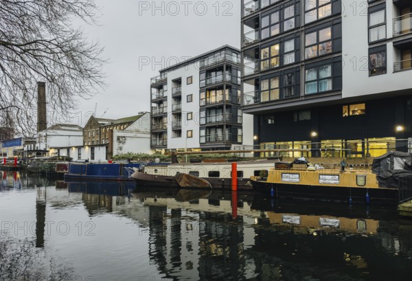 A serene London canal scene showcasing modern apartments alongside moored boats. The calm water reflects the architecture, creating an urban landscape blend