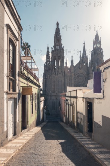 This image captures the stunning San Juan Bautista Church, located in Arucas, Gran Canaria, seen from a quaint, narrow street, lined with colorful traditional houses and cobbled pavement