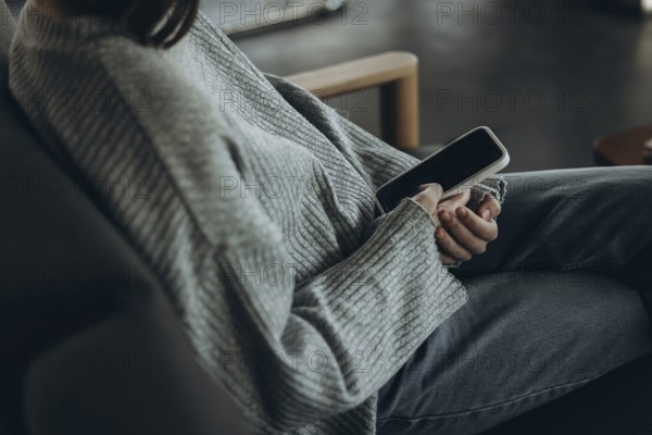 A person wearing a comfy sweater and jeans relaxes on a sofa, holding a smartphone. The setting exudes a cozy, casual home environment, perfect for unwinding