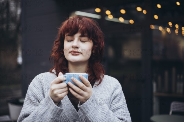 A young woman with red hair, in a gray sweater, enjoys a coffee on cafe terrace. String lights create a warm ambiance in the blurred background