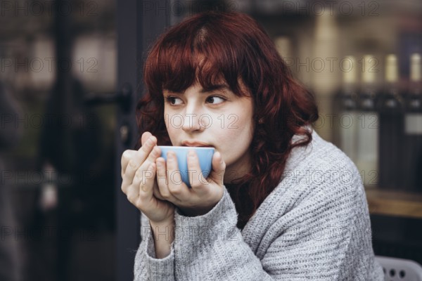 A young woman with red hair sips from a blue cup of coffee, wrapped in a cozy sweater. She gazes thoughtfully, creating a peaceful and introspective atmosphere