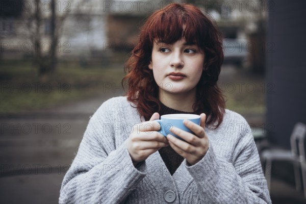 A young woman with red hair, wearing a grey sweater, holds a blue mug of coffee in an outdoor urban setting. She appears thoughtful, with a blurred background of buildings and trees on cafe terrace