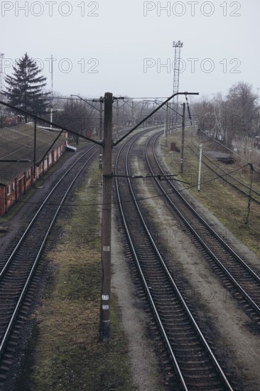 Overhead view of curved railway tracks in a misty, overcast setting. The tracks run alongside a weathered building, surrounded by leafless trees and a gray sky