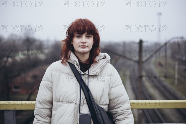 Young woman with red hair in a winter coat, standing on a bridge overlooking railway tracks in a day of travel by train. The cloudy sky and bare trees create a serene, moody atmosphere