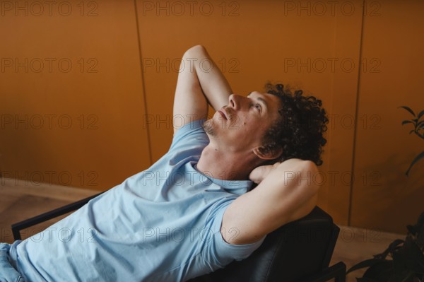A curly-haired man leans back in a chair with his hands behind his head, gazing upward. The warm orange wall and casual blue t-shirt create a relaxed, thoughtful atmosphere