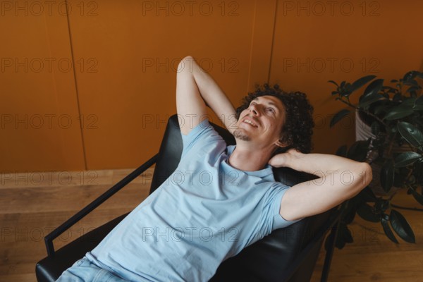 A curly-haired young man in a light blue shirt, lounging back in a modern chair with an orange accent wall and a green plant, exuding a calm and relaxed vibe