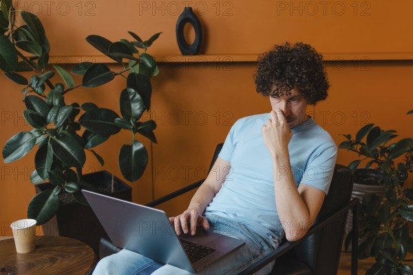 A curly-haired young man in a light blue shirt works on his laptop in a stylish room with plants and a warm orange wall, looking thoughtful and focused
