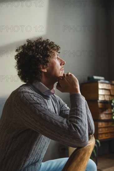 A contemplative man with curly hair sits by a window, resting his chin on his hands. Warm natural light highlights his face, creating a peaceful and introspective atmosphere