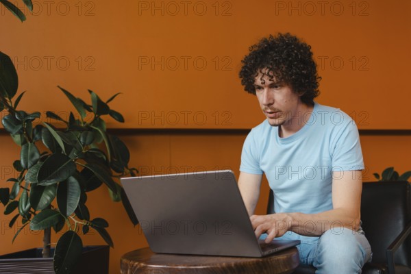A curly-haired young man focused on his laptop, sitting in a modern space with orange walls and greenery. Casual attire, relaxed atmosphere, productivity theme