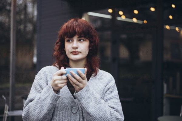 A young woman with red hair, wearing a gray cardigan, holding a coffee cup and gazing thoughtfully in a cozy cafe setting, surrounded by soft lighting and a warm atmosphere