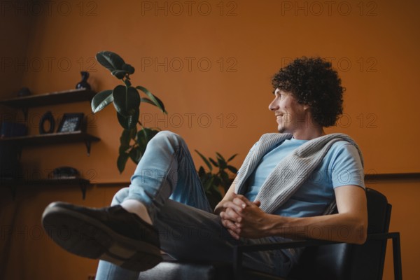 A curly-haired young man sits comfortably in a modern living room with a warm orange background, wearing casual attire. The setting exudes a cozy and relaxed atmosphere