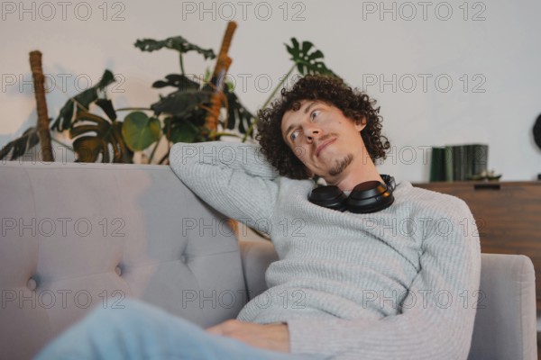 A curly-haired young man with casual attire, relaxing on a sofa with headphones around his neck. The serene setting features indoor plants in the background