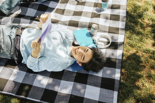 Friends lies on a checkered picnic blanket, enjoying a sunny day with headphones, papers, and coffee. The scene captures a blend of leisure and happiness